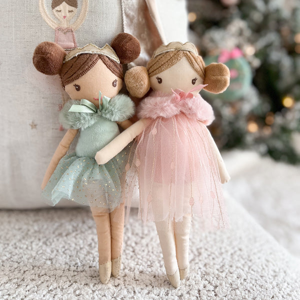 Two ballerina dolls in tulle dresses, one green and one pink, stand on a white textured surface.
