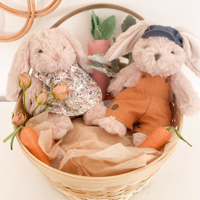 Two plush bunnies in a basket with carrots and flowers. One bunny wears a floral dress, the other wears overalls and a cap.