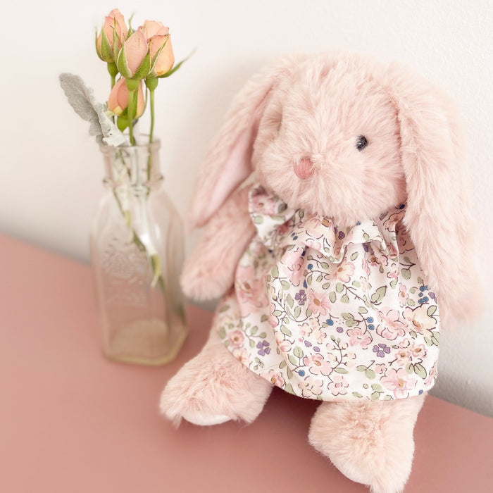 Close-up of a pink plush bunny wearing a floral dress, posed next to a small vase of pink roses.