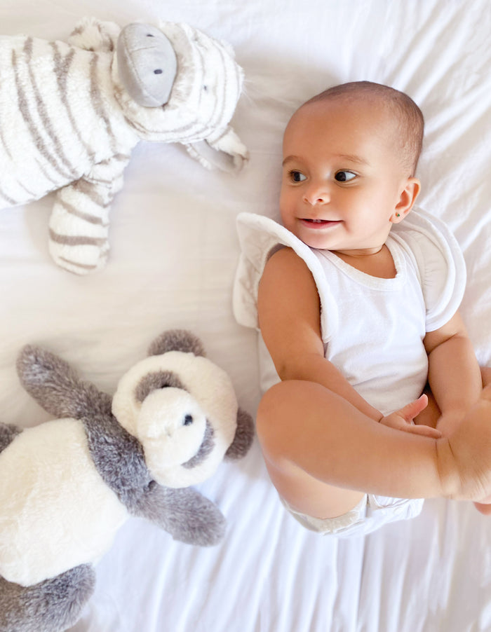 Overhead shot of baby in white onesie lying on white sheets with plush zebra and panda toys.