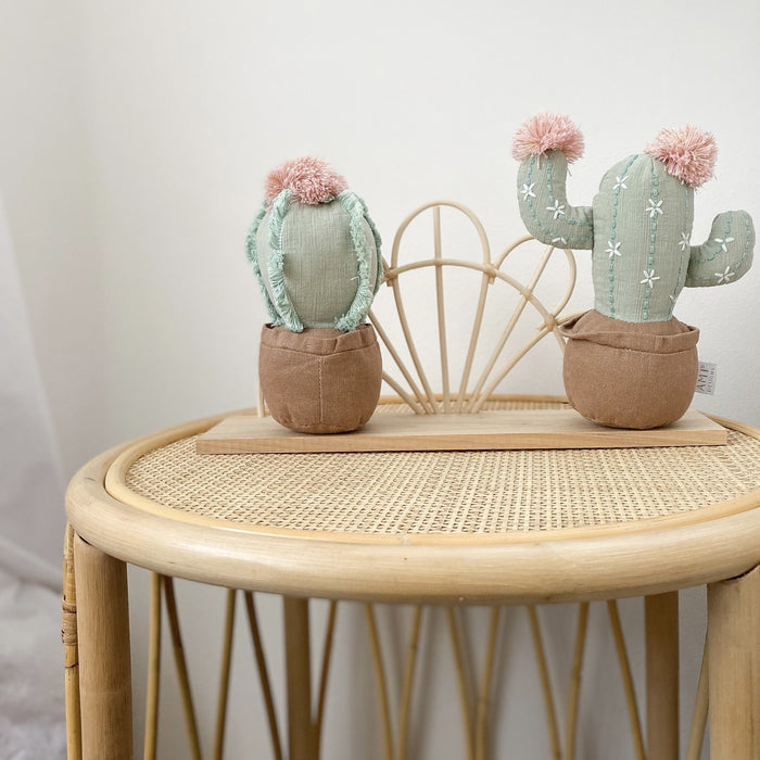 Two fabric cacti with pink pompoms in brown pots on a wooden tray, resting on a round wicker table.