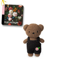 Crocheted brown teddy bear wearing black overalls with a pink flower embellishment, alongside a small Rijksmuseum tag featuring a floral still life.