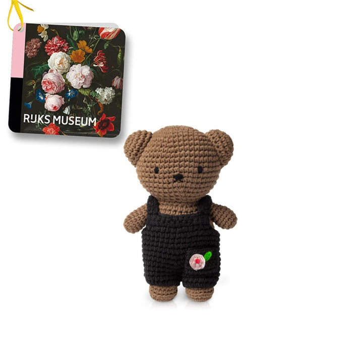 Crocheted brown teddy bear wearing black overalls with a pink flower embellishment, alongside a small Rijksmuseum tag featuring a floral still life.