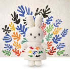 Crocheted Miffy toy in white dress with colorful leaf accents, posing against a backdrop of Henri Matisse-inspired cutouts.