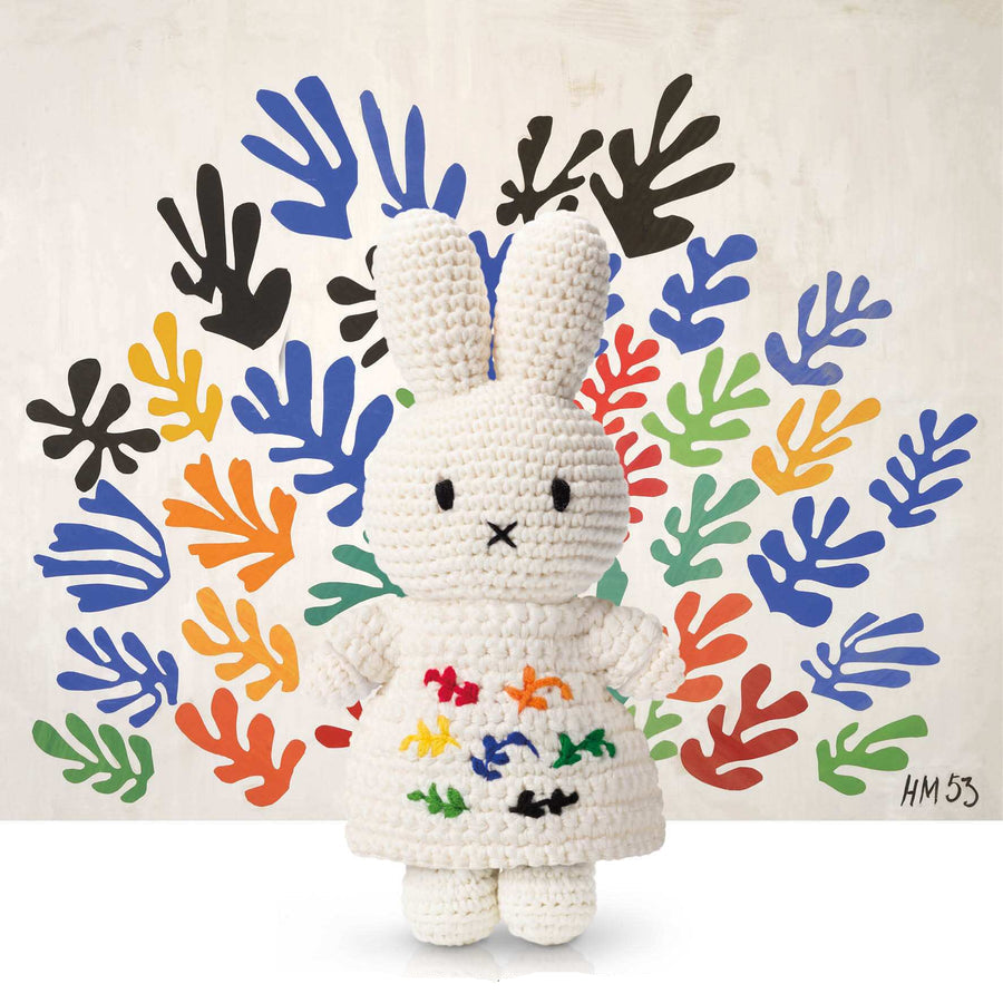 Crocheted Miffy toy in white dress with colorful leaf accents, posing against a backdrop of Henri Matisse-inspired cutouts.