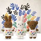 Three crocheted stuffed animals: a brown bunny, a white bunny, and a brown teddy bear, all wearing white outfits with colorful embroidery. They stand in front of a backdrop with colorful leaf-like shapes.