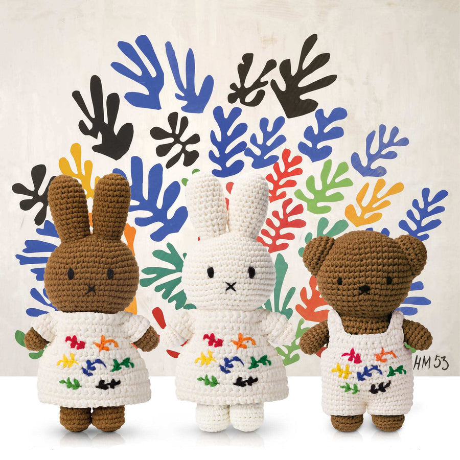 Three crocheted stuffed animals: a brown bunny, a white bunny, and a brown teddy bear, all wearing white outfits with colorful embroidery. They stand in front of a backdrop with colorful leaf-like shapes.