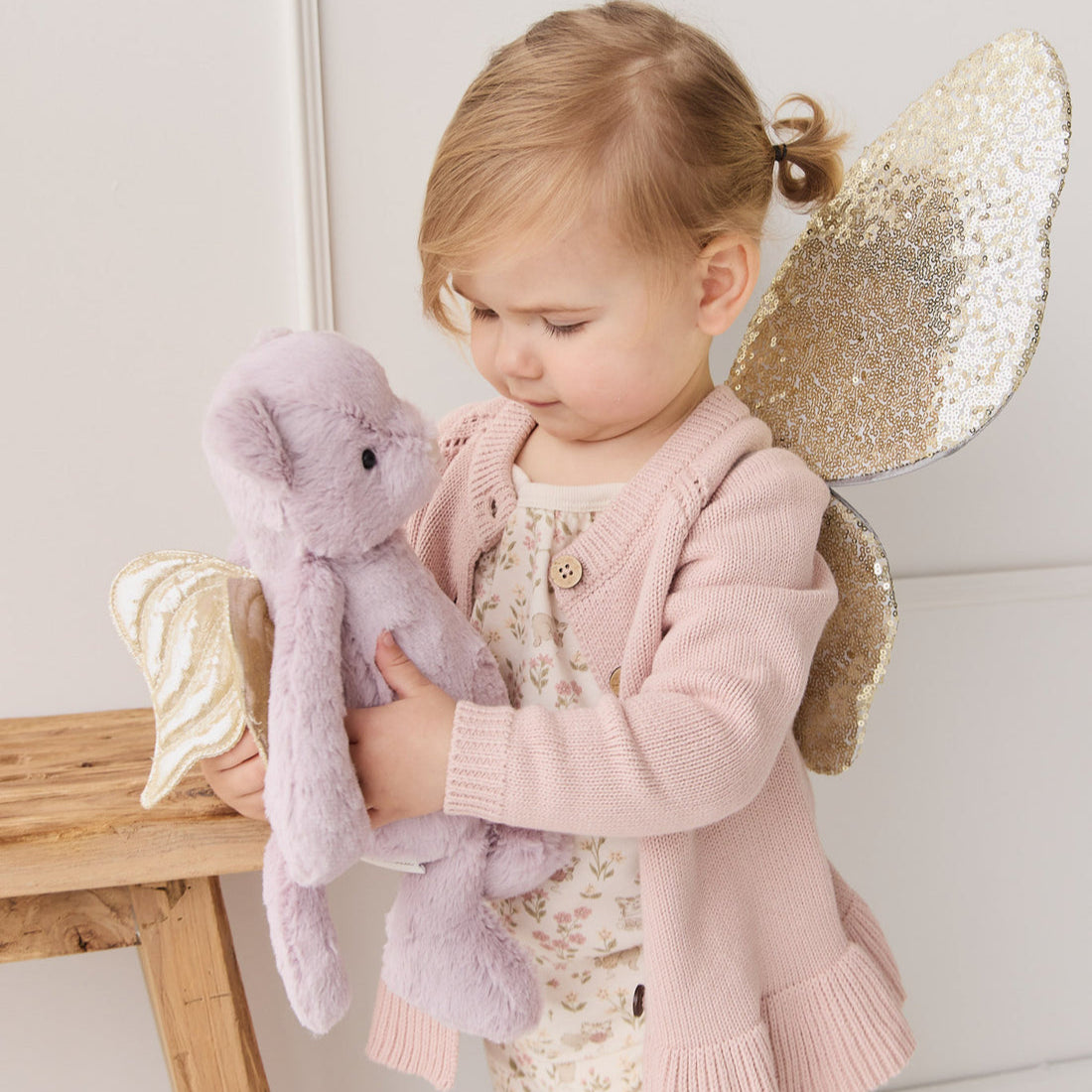 Young girl in pink cardigan holding lavender plush teddy bear with gold wings.