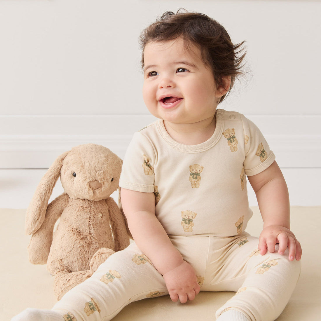 A baby sitting next to a stuffed bunny wearing teddy bear-printed pajamas.