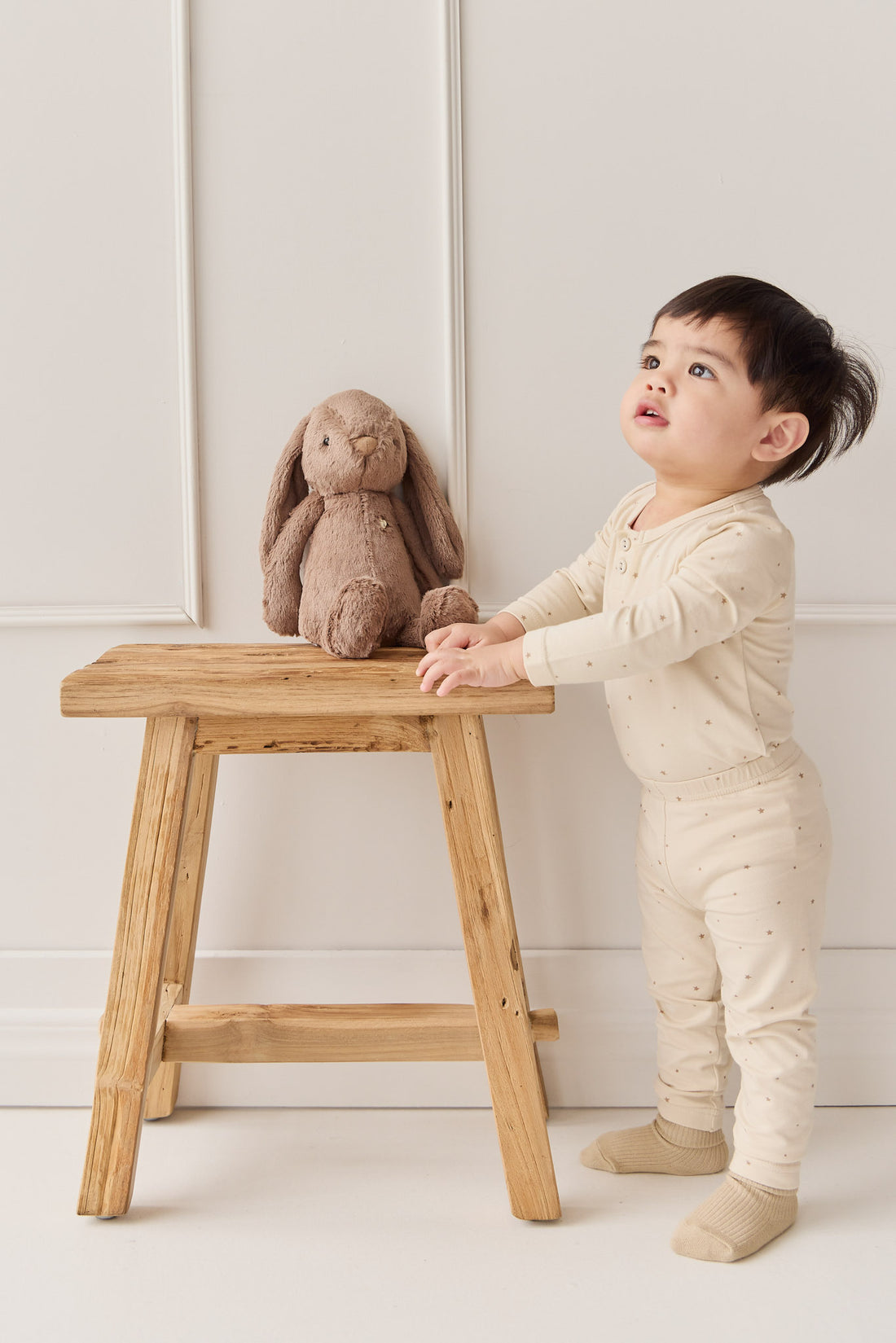 Toddler in beige pajamas with brown plush bunny on a wooden stool.