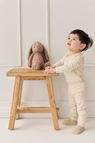 Toddler in beige pajamas with brown plush bunny on a wooden stool.