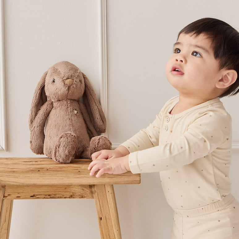 Toddler in beige pajamas with brown plush bunny on a wooden stool.