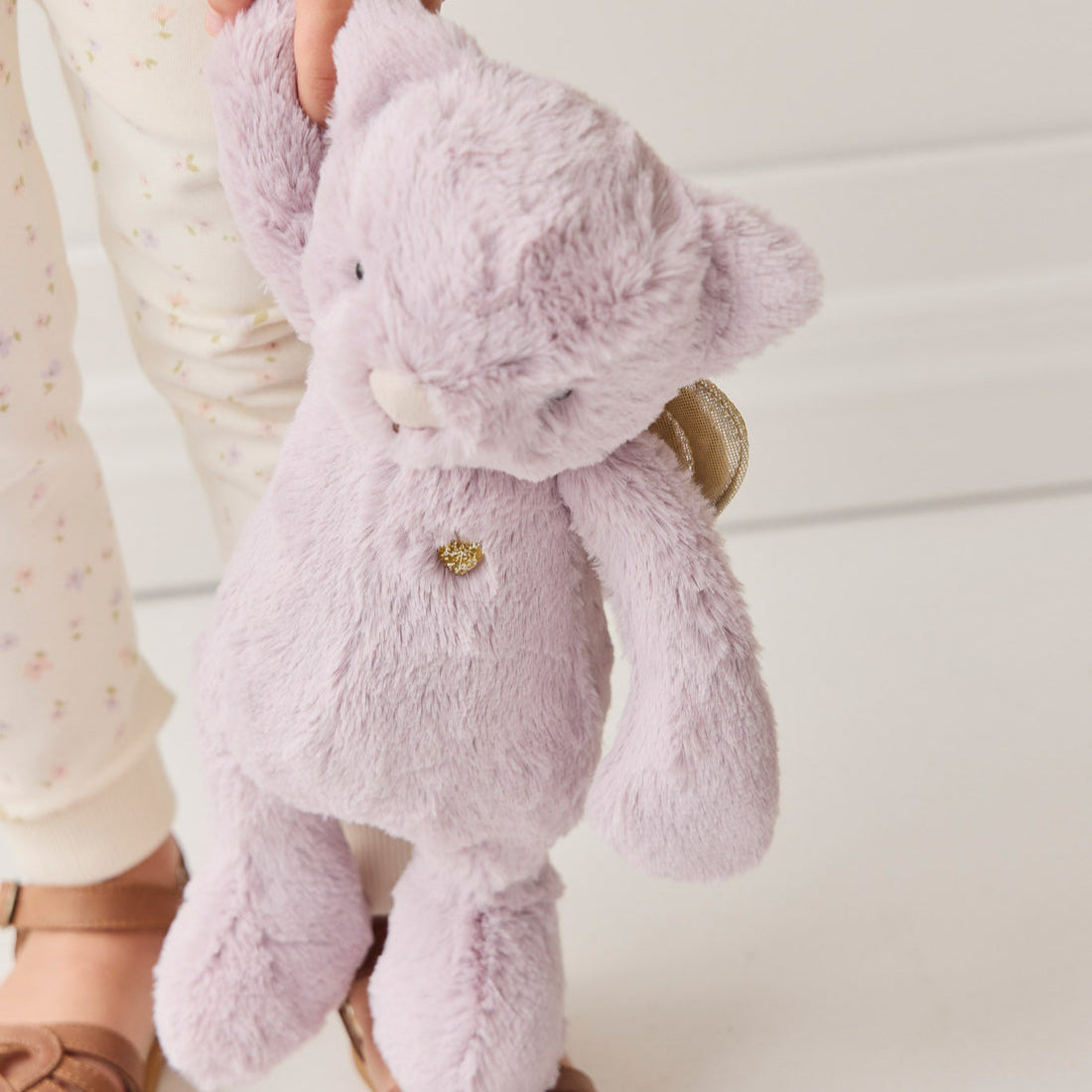Child holding a lavender stuffed bear with gold wings and a gold star on its chest, wearing floral print pajamas and brown sandals.