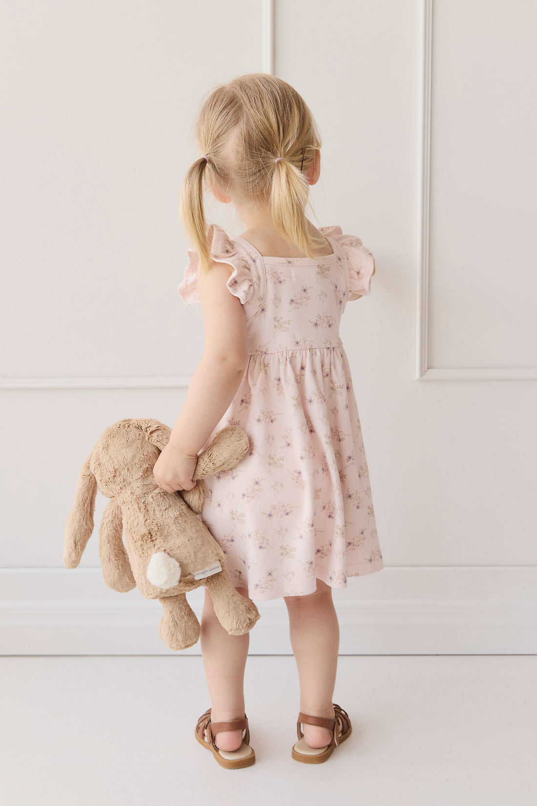 A little girl stands with her back to the viewer, wearing a pink floral dress and brown sandals, holding a plush bunny.