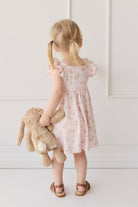 A little girl stands with her back to the viewer, wearing a pink floral dress and brown sandals, holding a plush bunny.