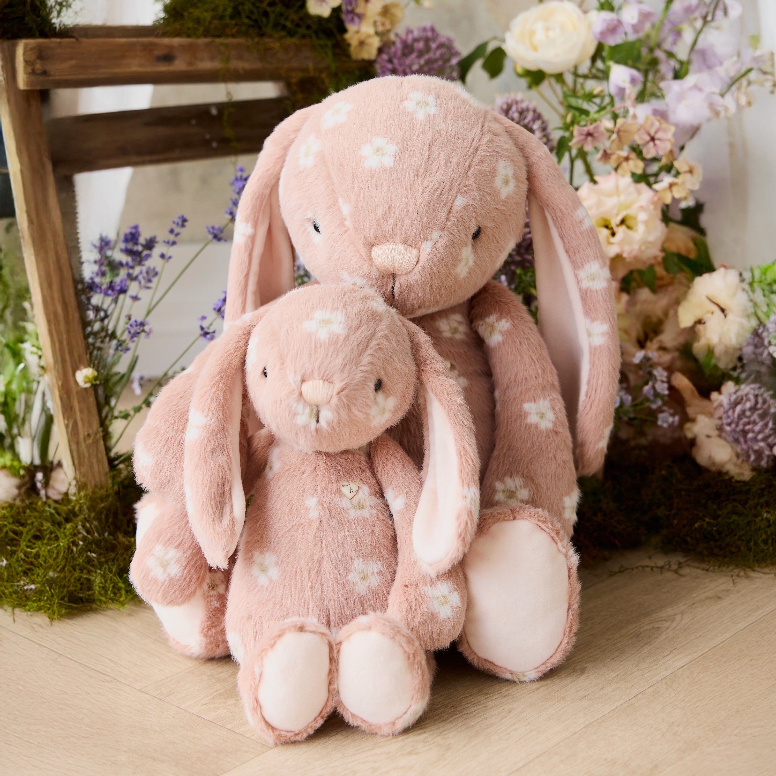 Two pink plush bunny toys with white floral patterns on a wooden surface with flowers in the background.