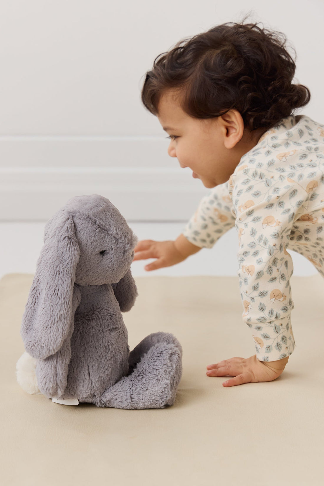 A baby in a patterned onesie reaches towards a gray stuffed bunny.