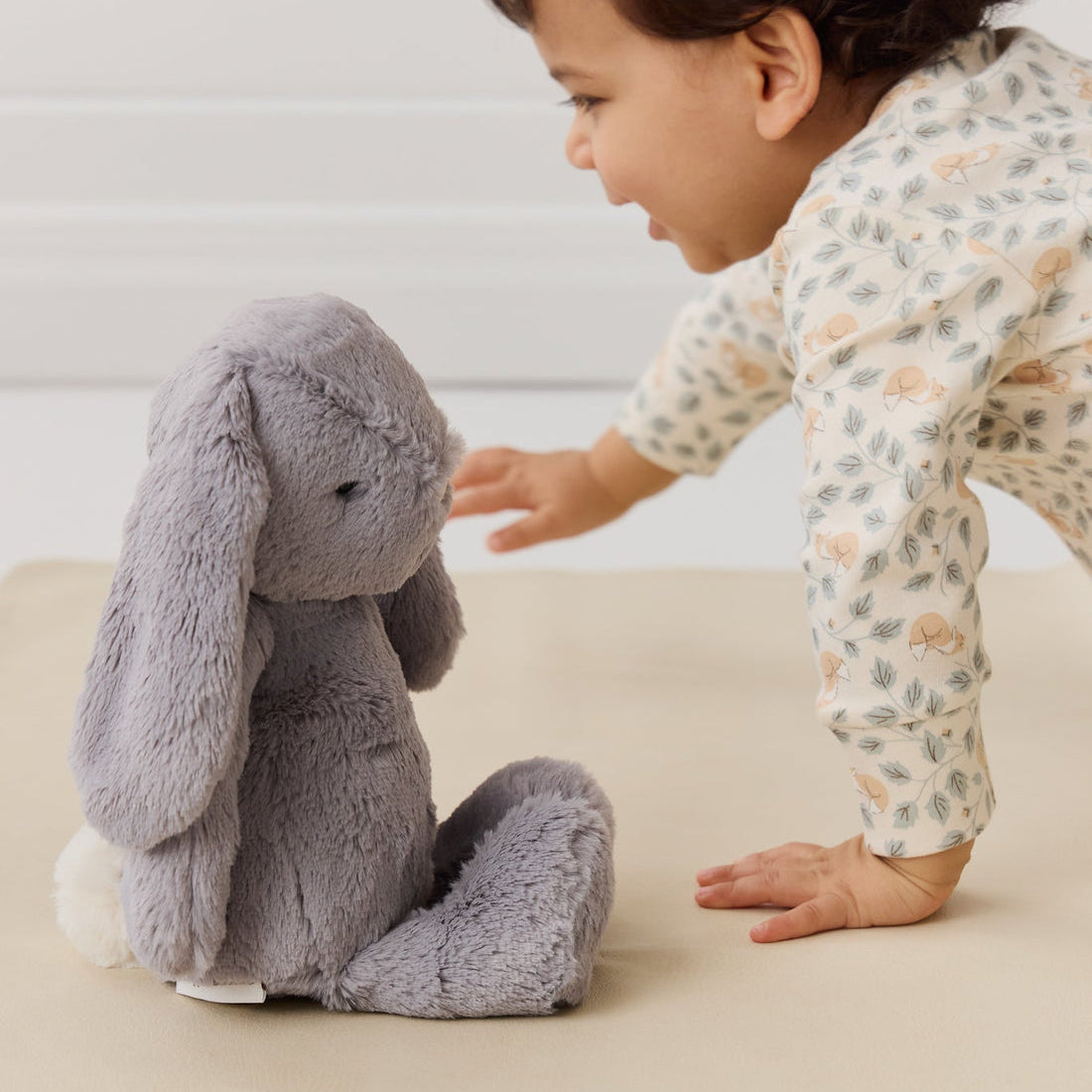 A baby in a patterned onesie reaches towards a gray stuffed bunny.