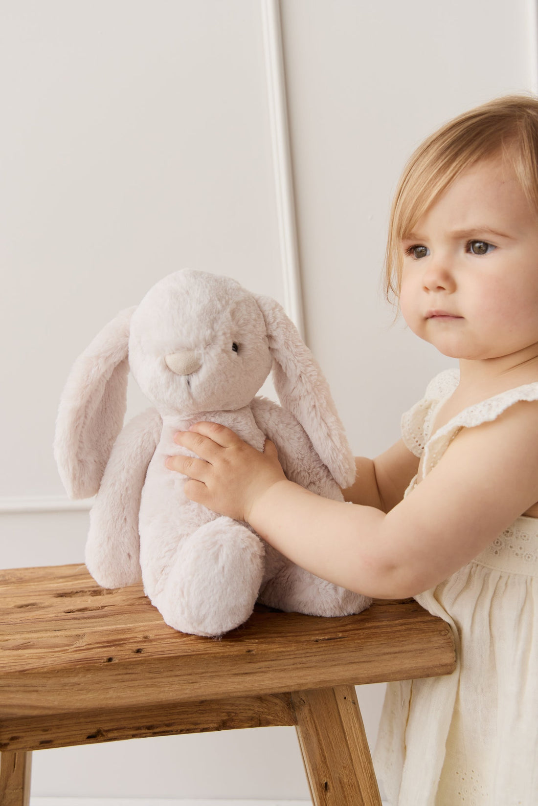 A toddler in a white eyelet dress sits next to a plush, light pink bunny on a wooden bench.