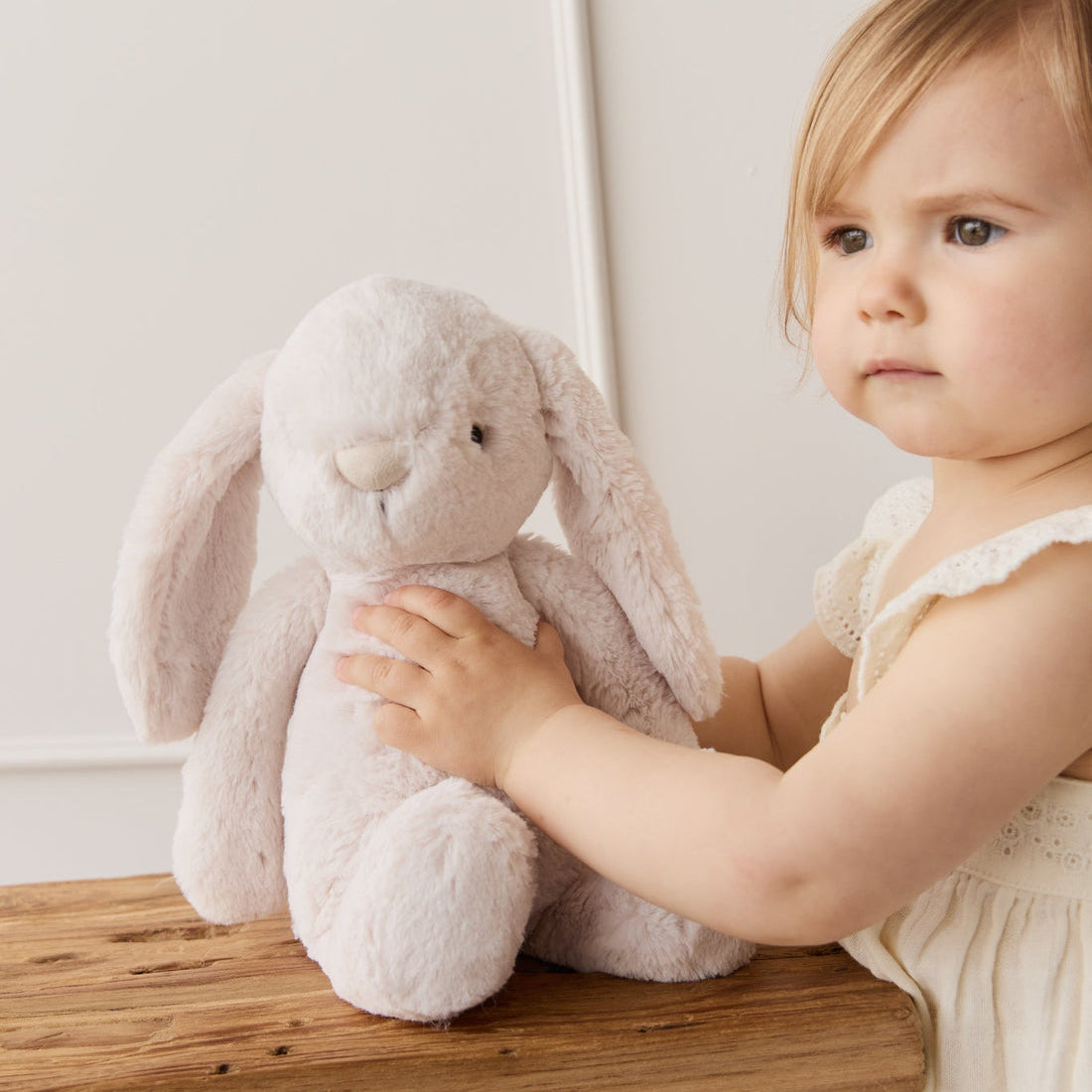 A toddler in a white eyelet dress sits next to a plush, light pink bunny on a wooden bench.