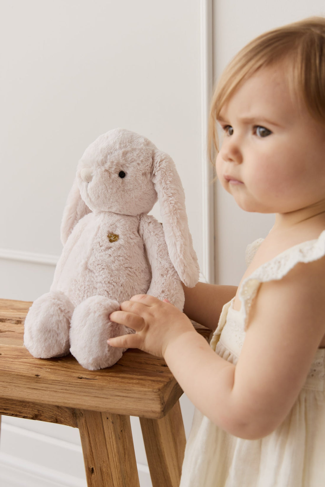 A toddler in a white dress touches a beige plush bunny on a wooden stool.