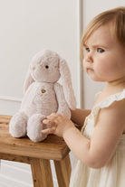 A toddler in a white dress touches a beige plush bunny on a wooden stool.