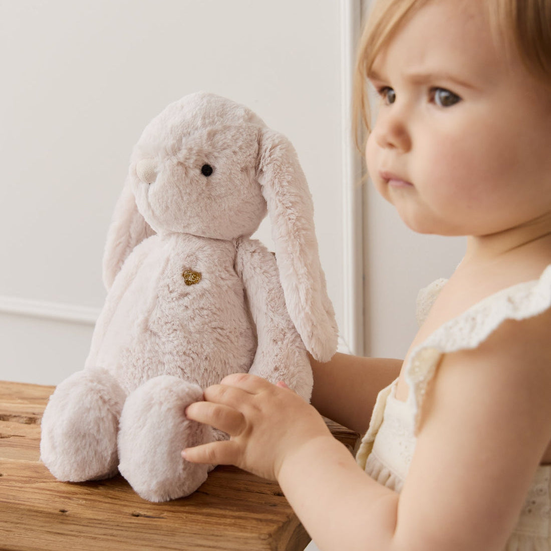 A toddler in a white dress touches a beige plush bunny on a wooden stool.
