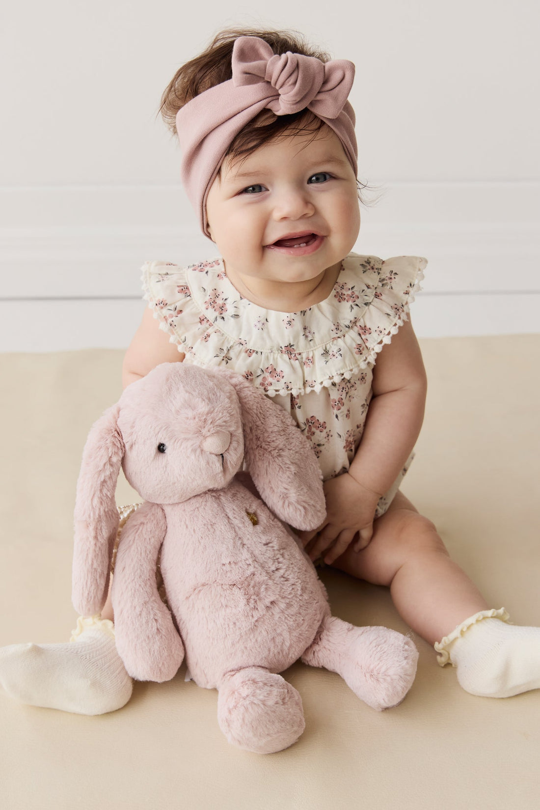 A baby girl sits with a pink stuffed bunny toy, smiling and wearing a pink headband and a floral print ruffled-neck romper.
