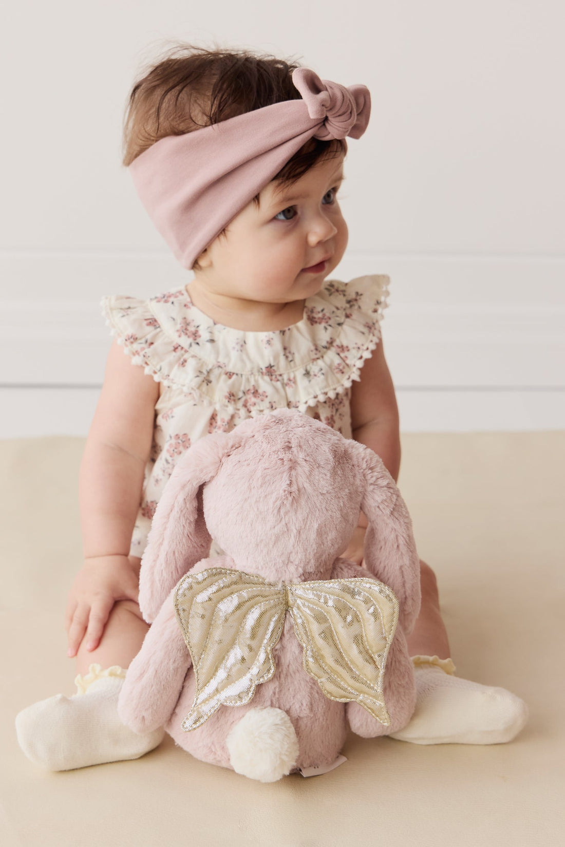 Baby with pink headband and floral romper sitting with pink bunny plush with gold wings.