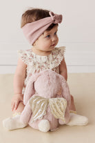 Baby with pink headband and floral romper sitting with pink bunny plush with gold wings.