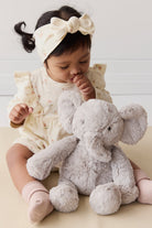 Baby with floral headband and outfit sitting with a plush gray elephant toy.