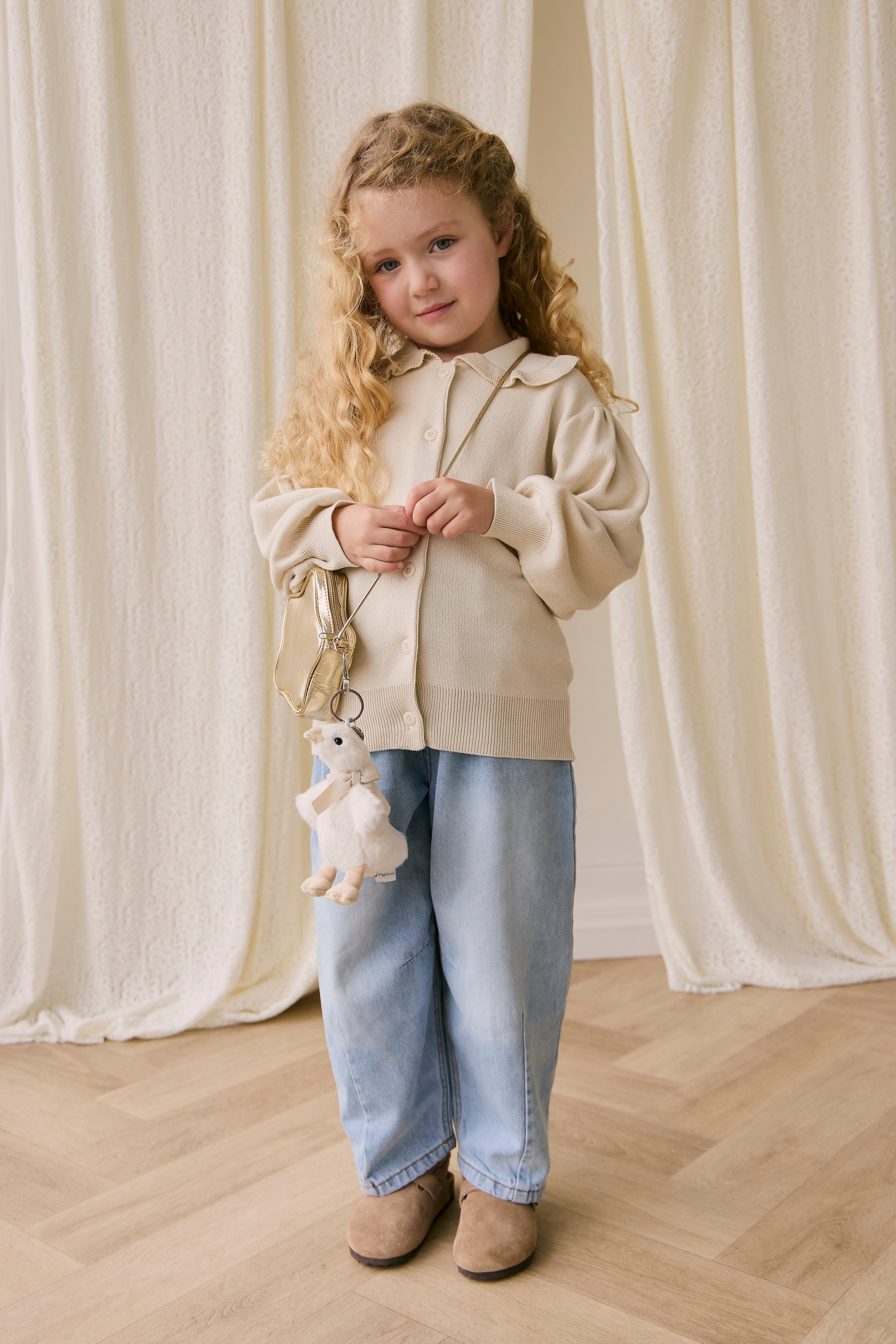 Young girl in a beige jacket and blue jeans holding a white teddy bear against a white curtain background.