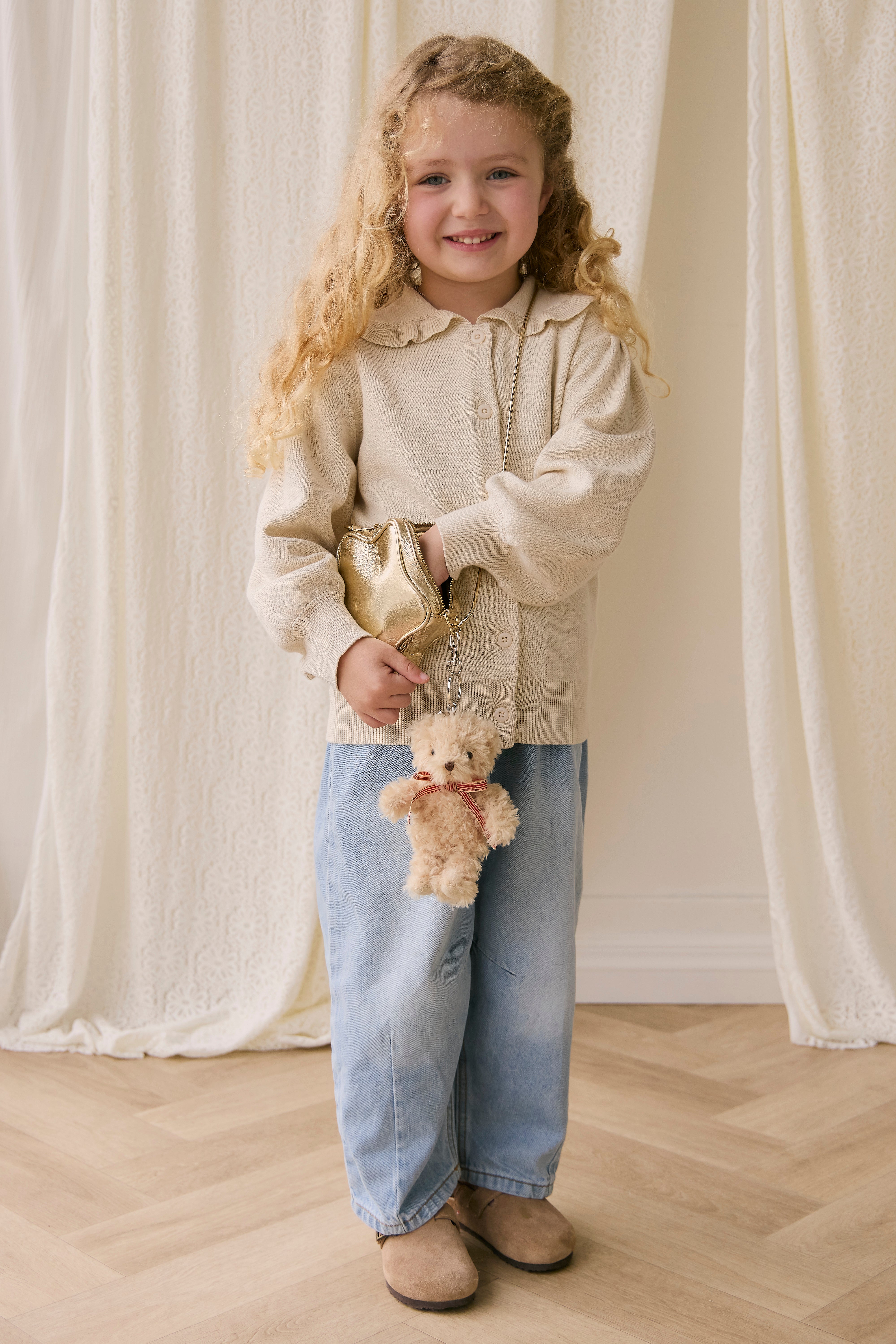 Young girl holding a teddy bear and a gold bag against a white curtain background