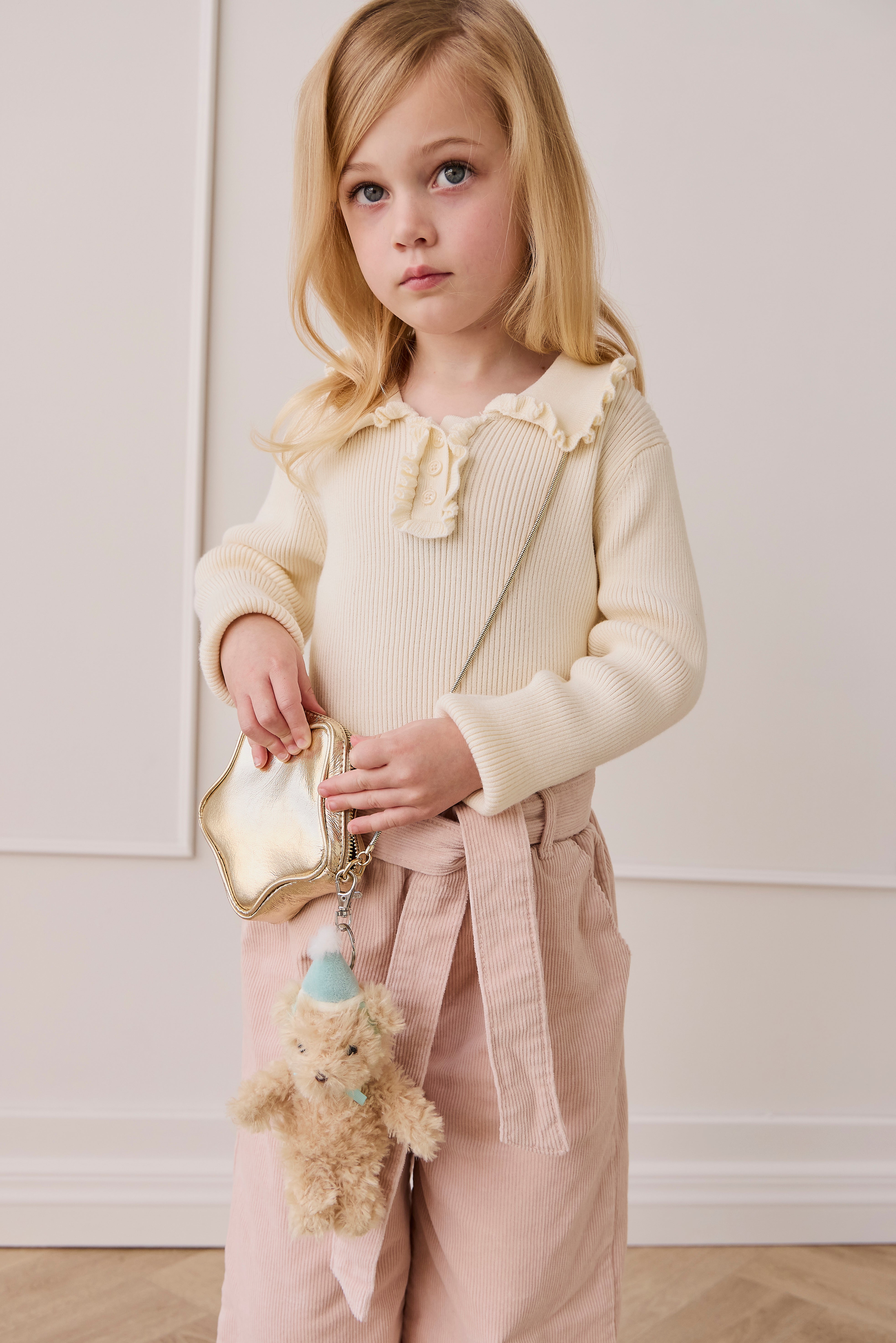 Young girl in a cream sweater and pink pants holding a gold handbag with a teddy bear charm.
