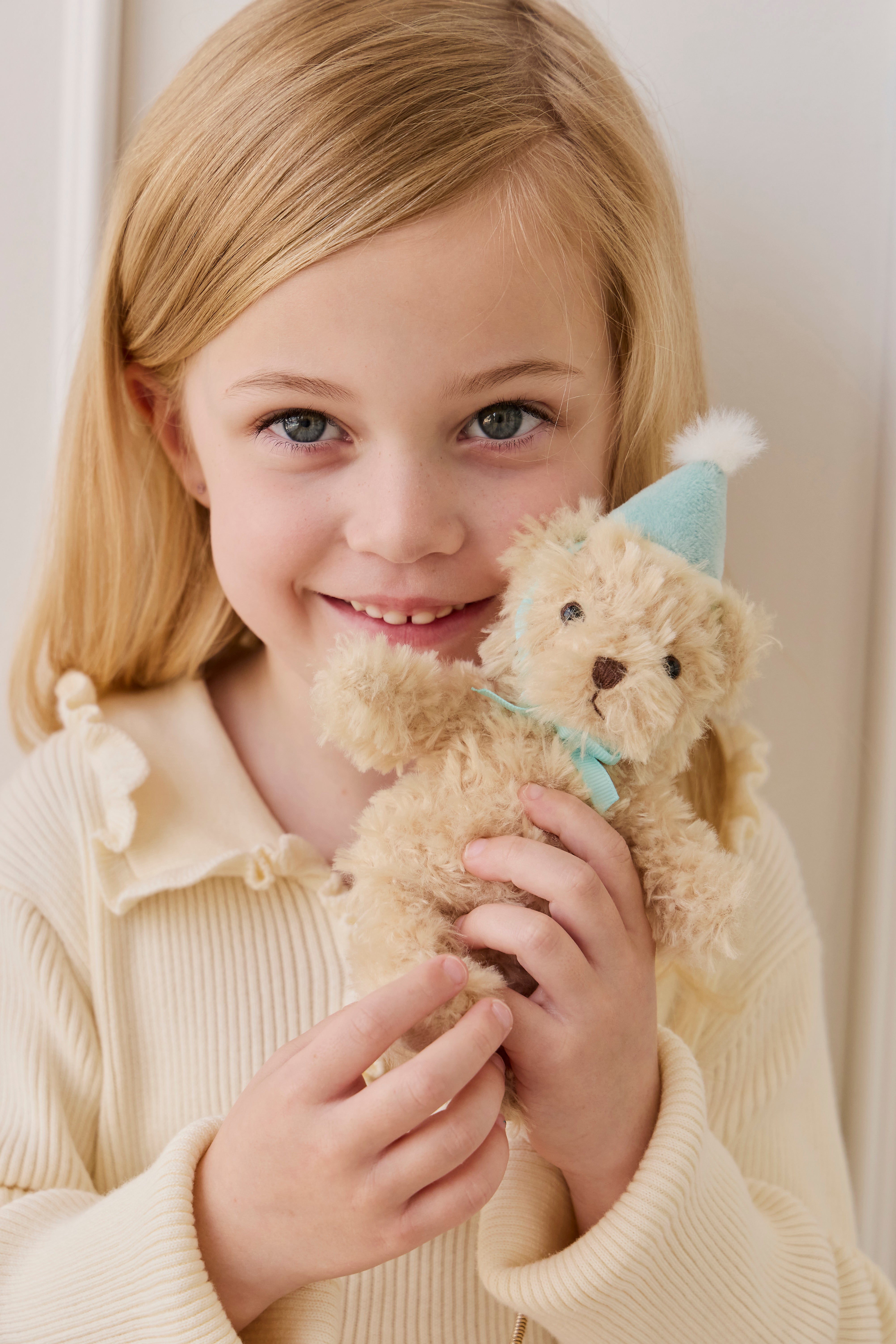 Young girl holding a teddy bear with a party hat against a plain background