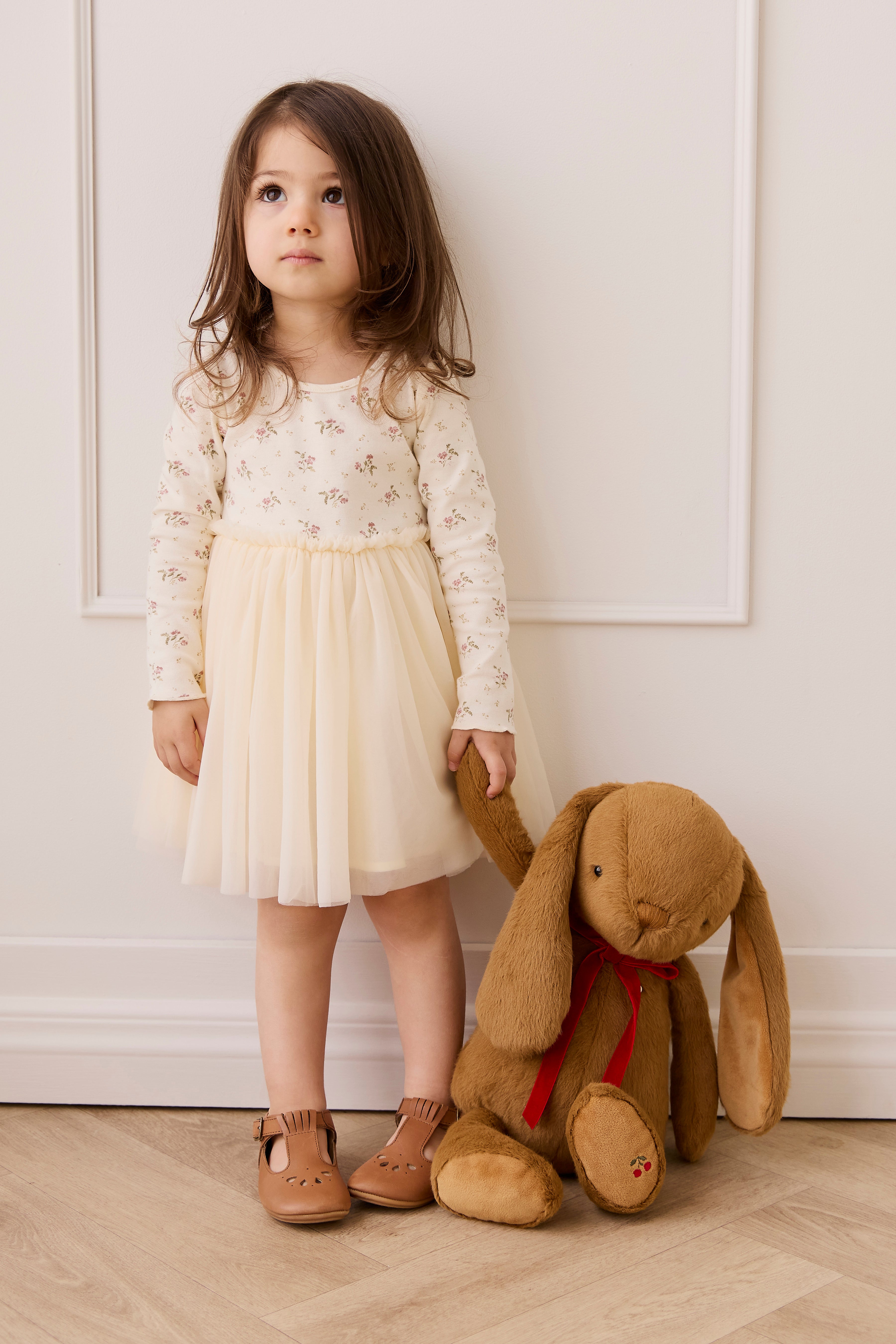 Young girl in a white dress holding a brown teddy bear against a white wall.