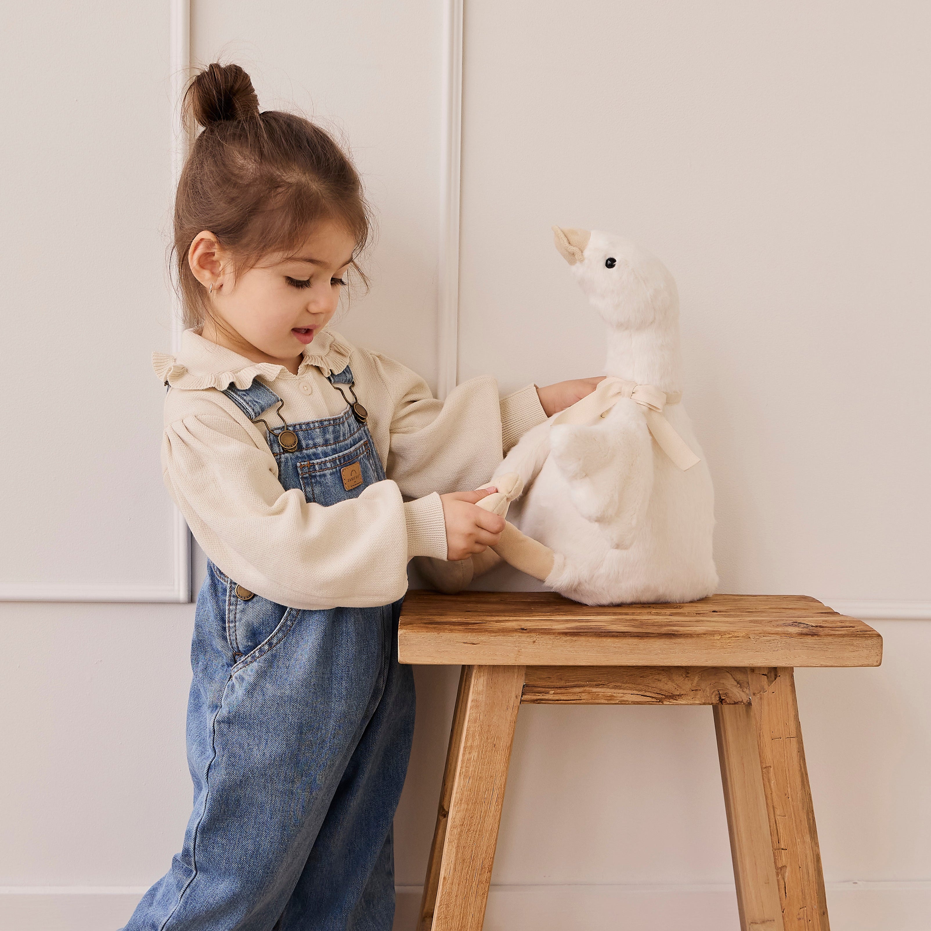 Child in denim overalls interacting with a plush toy on a wooden stool against a white wall.