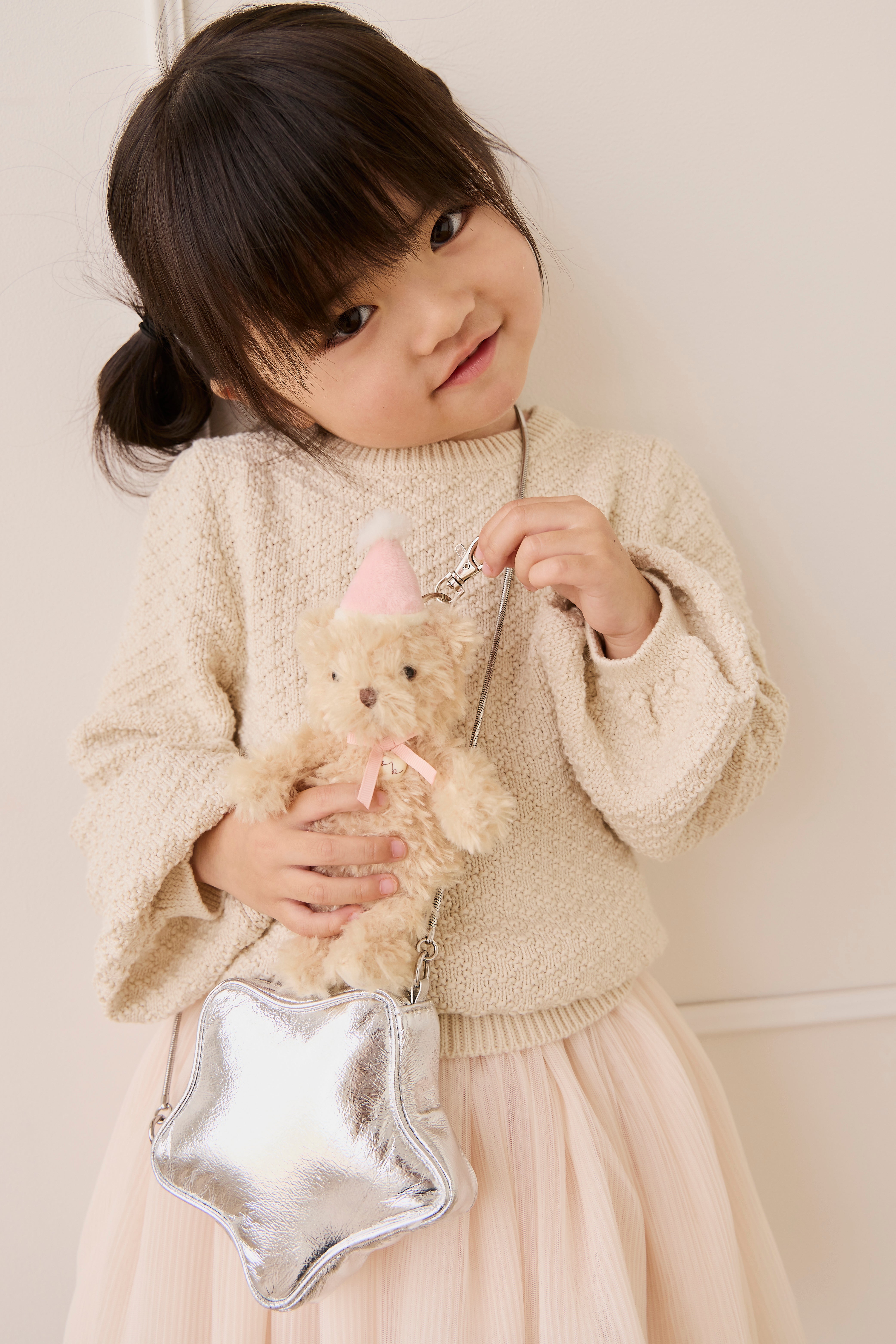Young girl holding a teddy bear and a silver star-shaped bag against a white background