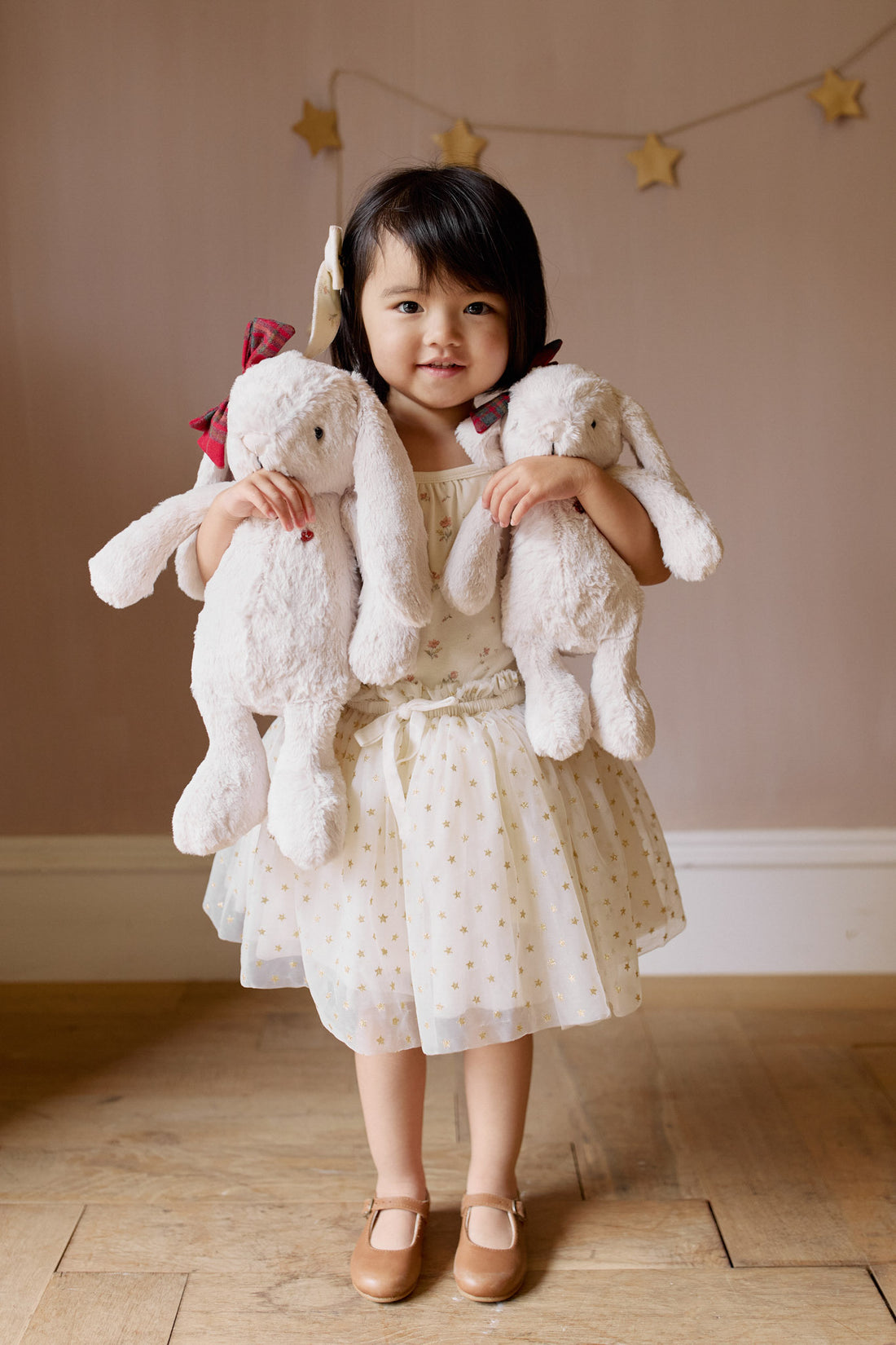 Toddler girl holding two white plush bunny dolls, wearing floral and star dress and brown Mary Jane shoes, standing indoors.