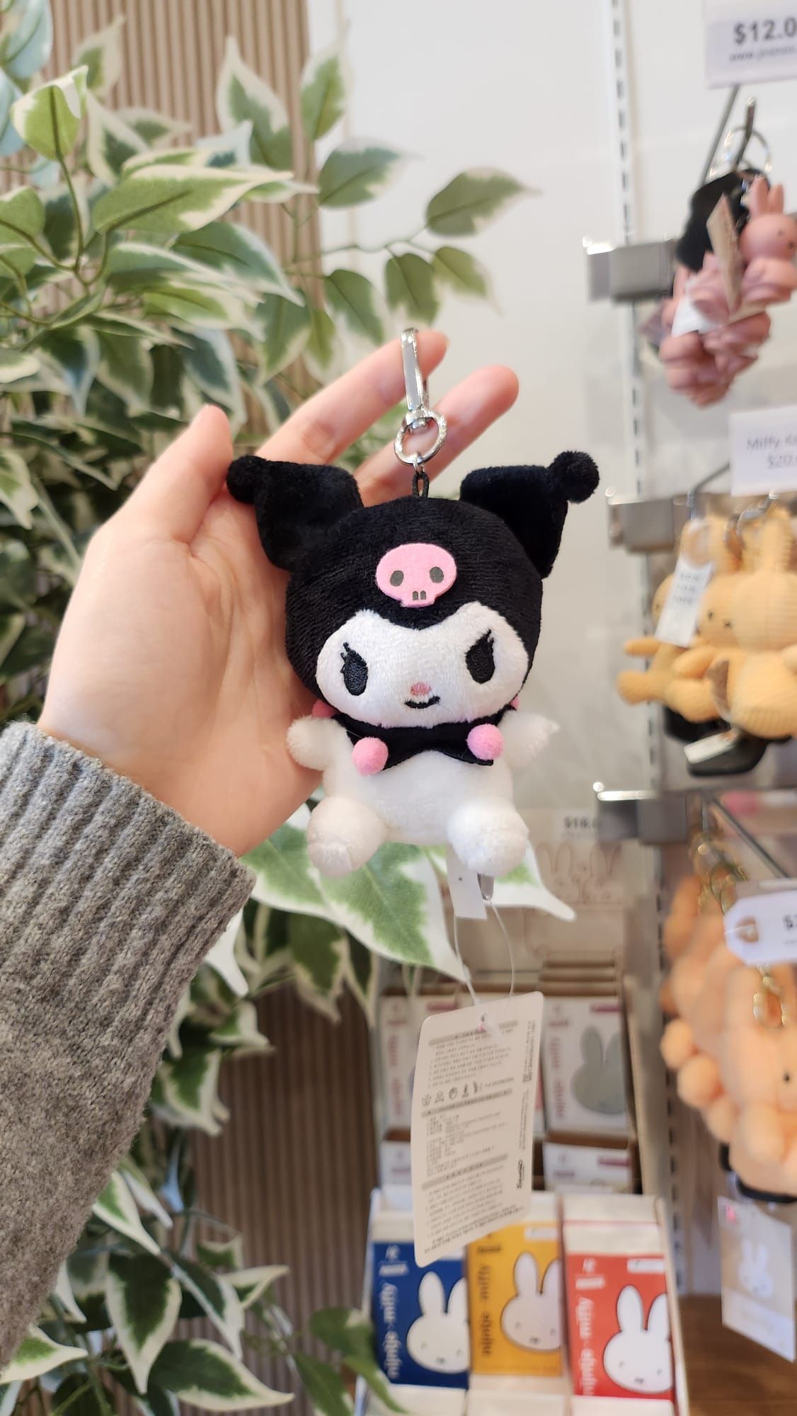 Hand holding a black and white Kuromi plush keychain with a pink skull on its head, with shelves of Miffy merchandise in the background.