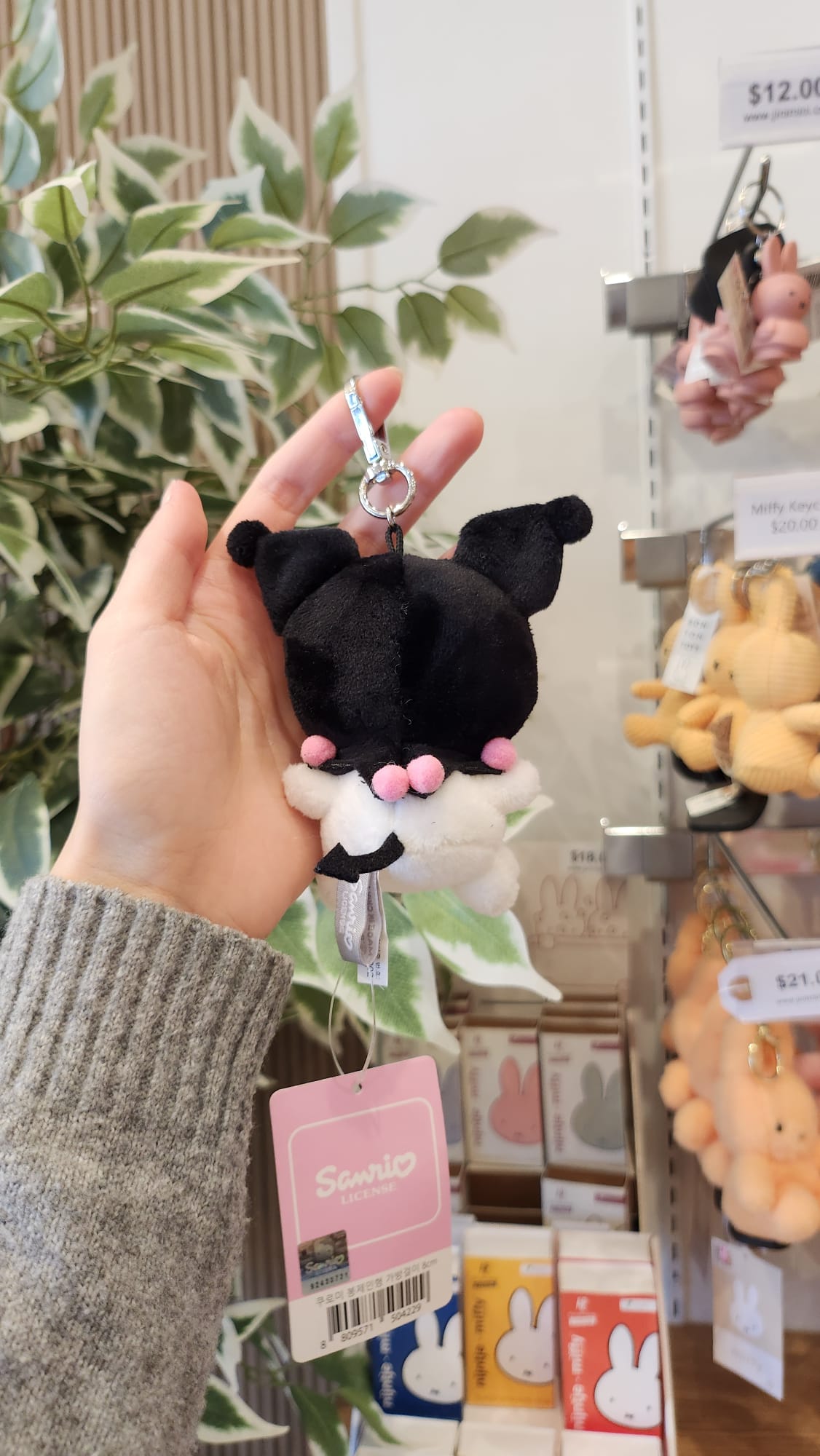 A hand holds a Kuromi keychain plush with a pink Sanrio tag, near shelves of Miffy merchandise.