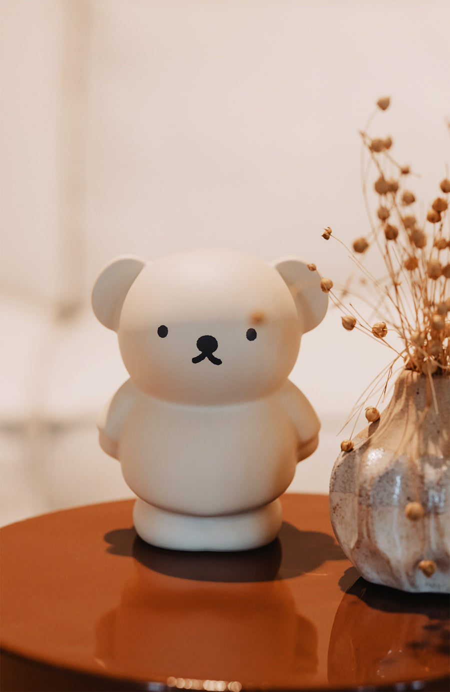 Beige bear-shaped toy and speckled vase with dried flowers on glossy brown surface.