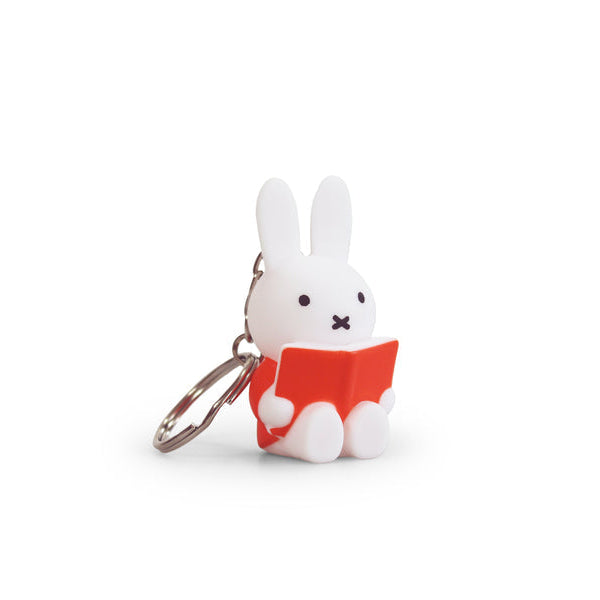 A white, Miffy, keychain, sitting while reading a red book.