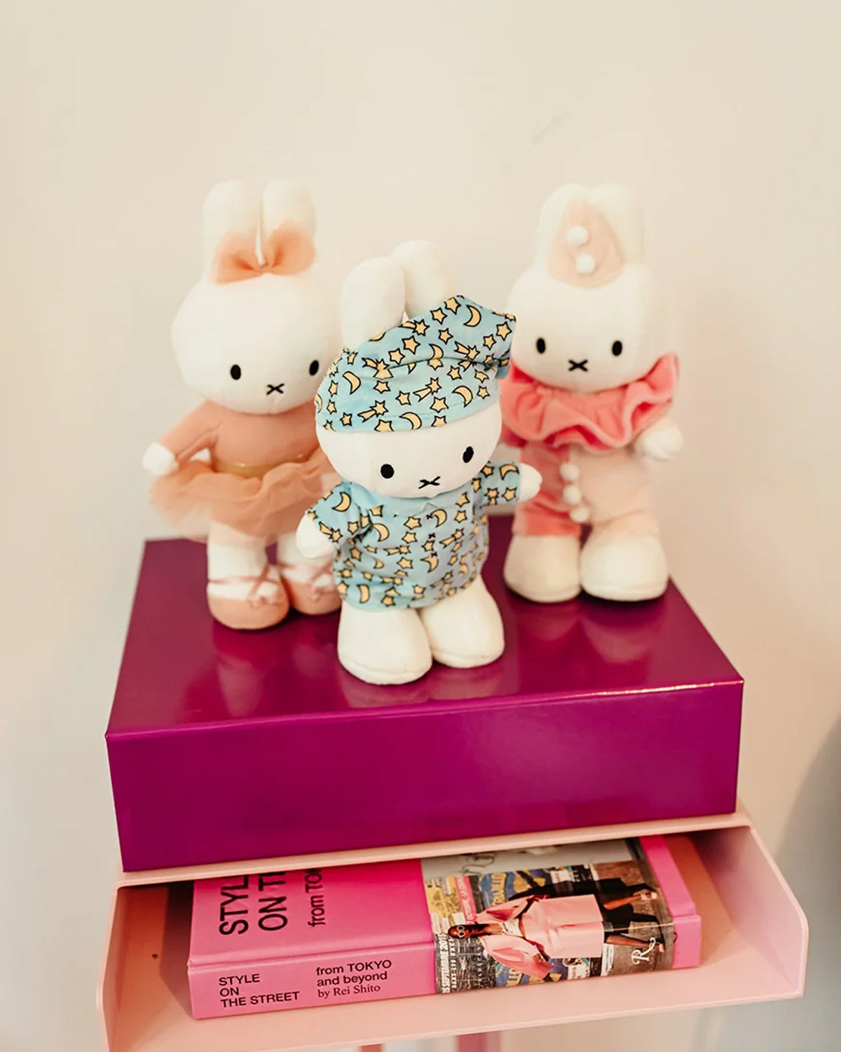 Three Miffy dolls wearing different outfits stand on a purple box resting on a shelf with a pink book.