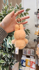 A hand holds a fuzzy, peach-colored Miffy bunny keychain with a gold keyring and "nijntje miffy" tags. Behind the hand is a green and white leafy plant and a display of Miffy products.