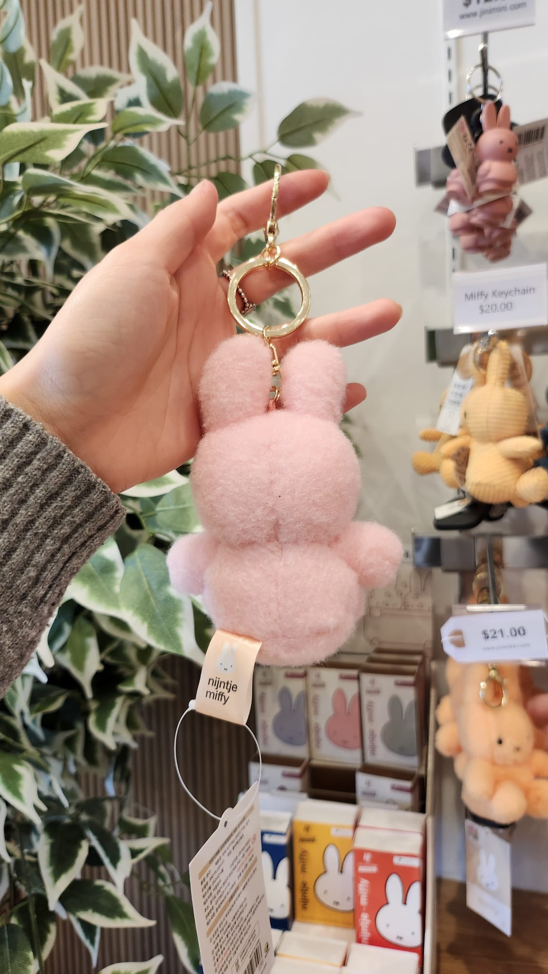 A pink Miffy keychain is held in hand. The keychain has a tag with the words "nijntje miffy." Other Miffy keychains in various colors and prices are visible on a display shelf. A leafy plant is in the background.