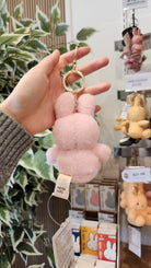 A pink Miffy keychain is held in hand. The keychain has a tag with the words "nijntje miffy." Other Miffy keychains in various colors and prices are visible on a display shelf. A leafy plant is in the background.