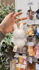 A hand holds a white, plush Miffy keychain, with Miffy-themed merchandise and other keychains displayed on a shelf in the background.