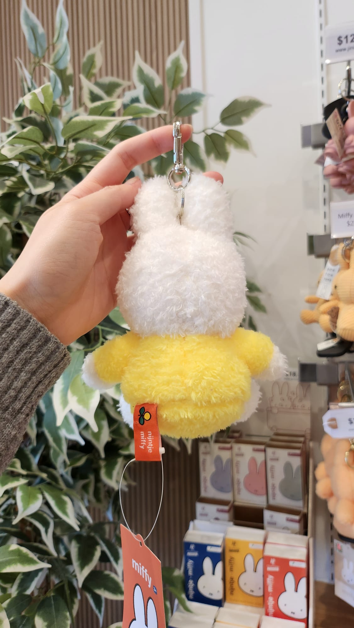 A person holding a Miffy keychain featuring a white fuzzy head and a yellow fuzzy body with an orange tag.