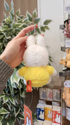 A person holding a Miffy keychain featuring a white fuzzy head and a yellow fuzzy body with an orange tag.